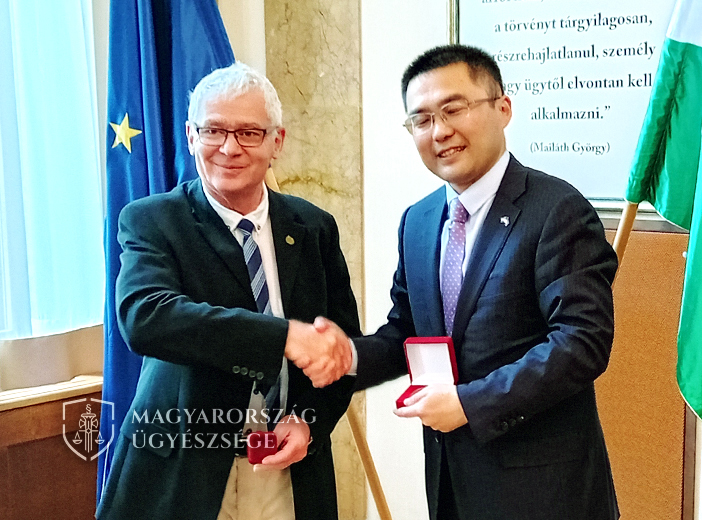A fotón dr. Polt Péter legfőbb ügyész kezet fog a magyar és az uniós zászló előtt a Kínai Népköztársaság ideiglenes ügyvivőjével.
